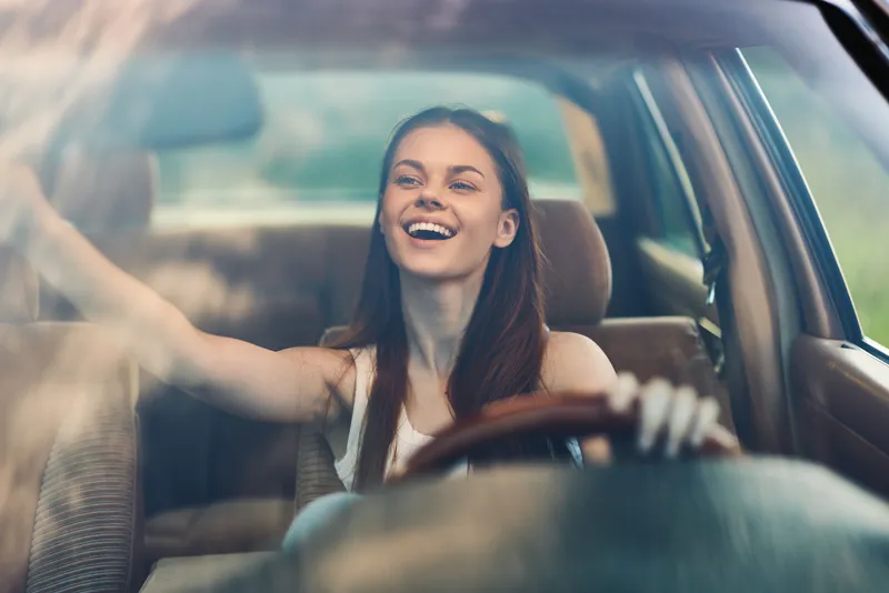 Young woman smiling while driving a car, enjoying freedom and adventure, bright daylight, casual outfit, appealing automotive interior, happiness, lifestyle image.