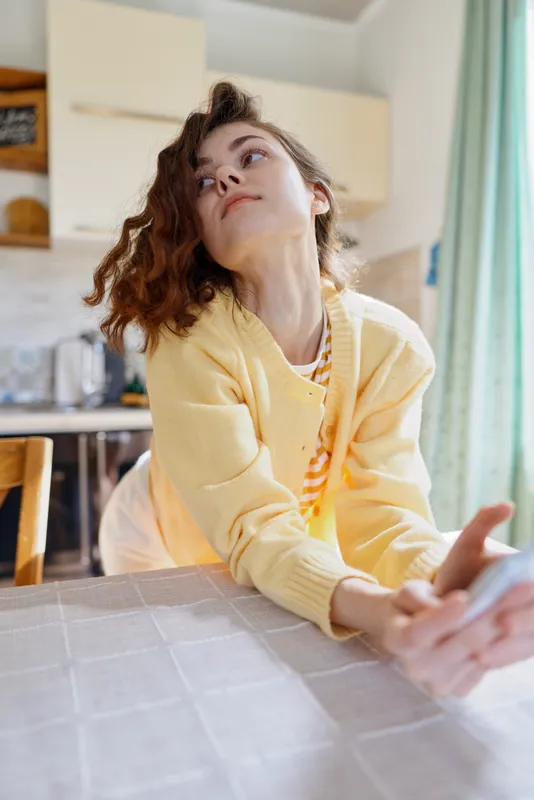 A stressed woman in a yellow cardigan leans on a kitchen table, looking upward in thought. A candid home lifestyle moment highlighting fatigue, resilience, and everyday mental load.