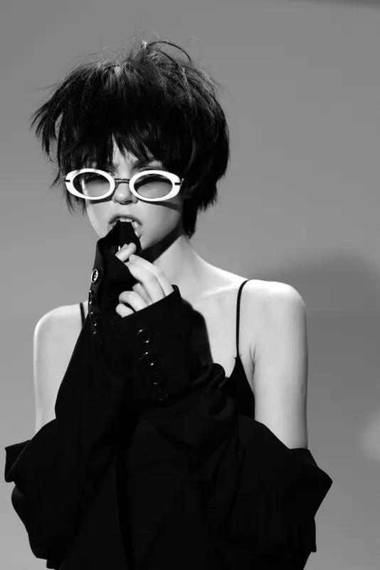 Black and white portrait of a young woman in white glasses wearing a black outfit with short messy hair.