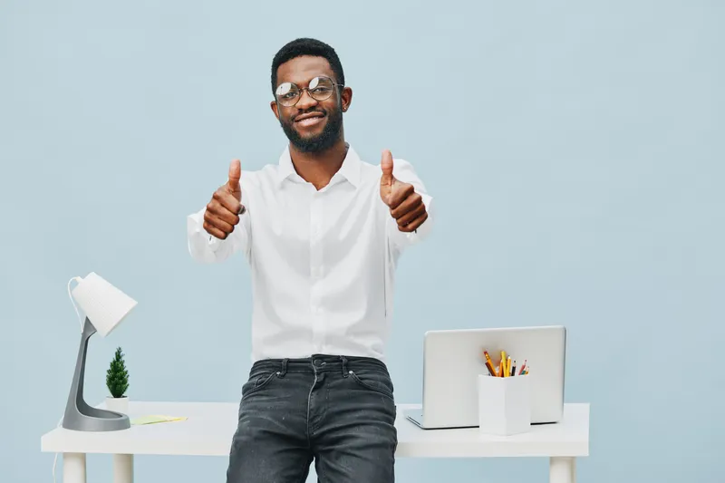 Smiling happy young man with glasses standing behind white office desk, giving thumbs up, wearing white shirt and black jeans, modern workspace with laptop, stationery, and small plant, cheerful busin