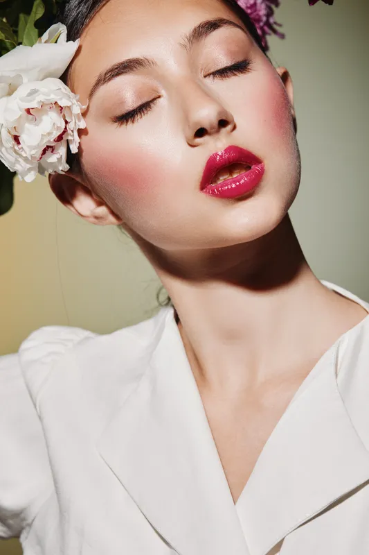 Close-up of a female beauty model with glossy red lips and smooth skin wearing pink and white floral headwear. Soft lighting emphasizes high fashion and creative makeup art concepts.