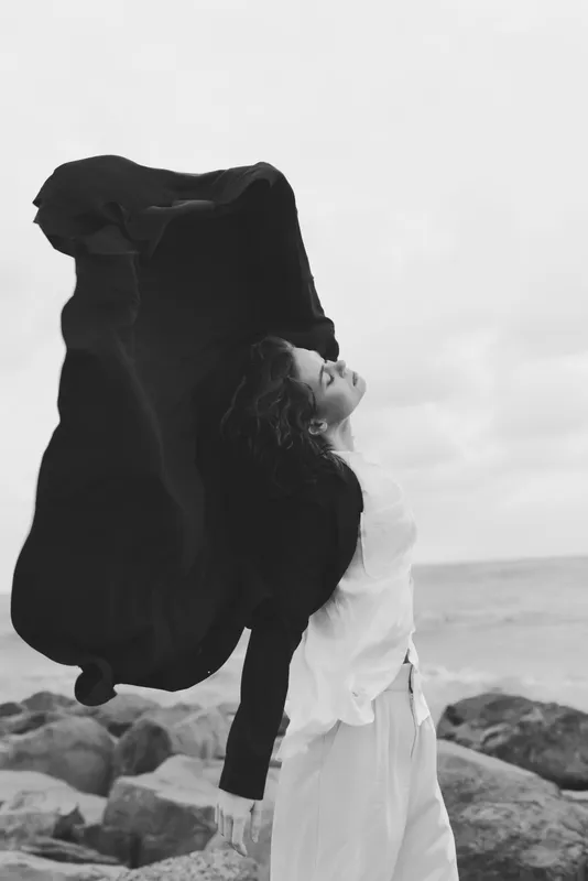 Woman on a windy beach lifts a flowing coat creating a dramatic silhouette against the sea and sky conveying freedom movement and minimalist fashion mood