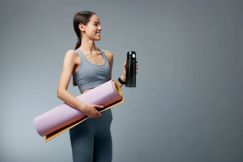 Fitness woman in athletic wear stands with a water bottle and rolled yoga mat, ready for a workout. Neutral backdrop highlights strength, balance and healthy lifestyle for gym or studio use.
