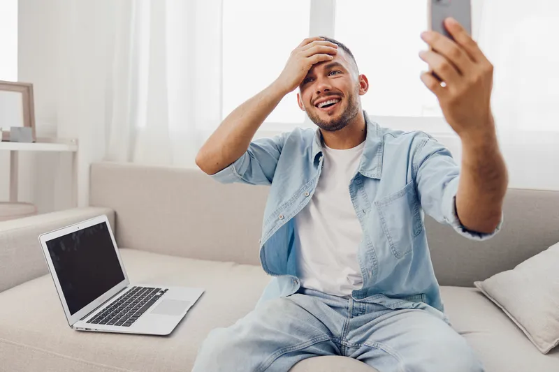 Happy man taking a selfie while sitting on a couch, expressing emotions related to mental health and work-life balance, with a laptop nearby.