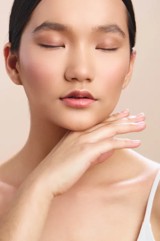 Asian woman with serene expression and soft makeup rests eyes closed in a neutral studio setting, conveying calm beauty, focus, and peaceful presence.