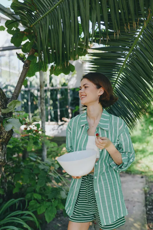 A young woman in a striped green shirt walks outdoors beneath palm fronds, smiling as she holds a white hat.