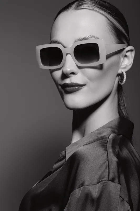 Stylish caucasian woman with fair skin wearing large sunglasses and earrings poses in silky blouse on dark background.