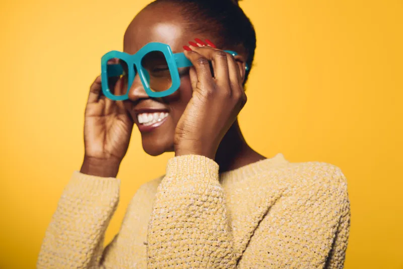 Smiling dark skinned African woman wearing oversized blue sunglasses and yellow sweater. Portrait on bright yellow background. Concept of fashion, style, happiness, and confident beauty.