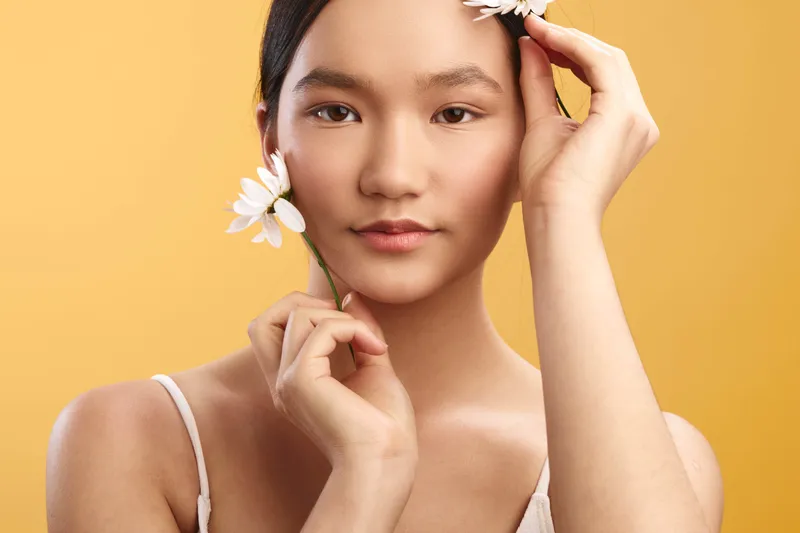 asian woman gazes softly at the camera, holding white flowers near her face against a warm background, conveying natural beauty, calm charm, and gentle elegance.