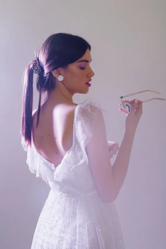 Elegant woman in a white dress with an open back holds a delicate necklace. Soft lighting highlights her beauty and fashion style in a minimalist studio setting.
