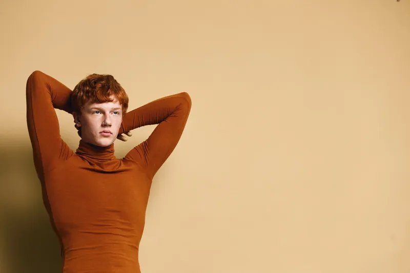 Young man with red hair and freckles posing with arms behind head wearing brown turtleneck against beige background studio portrait.