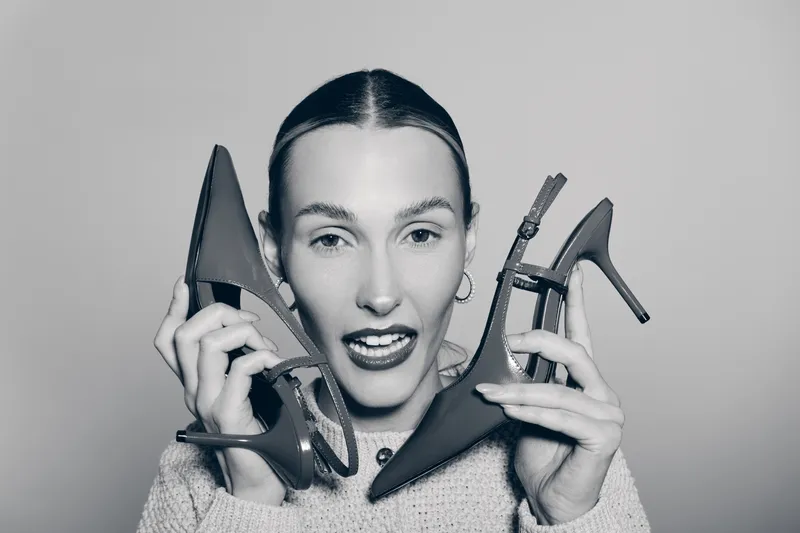 Young white woman with light skin holds two black high heel shoes near her face. Studio portrait on grey background with playful expression.