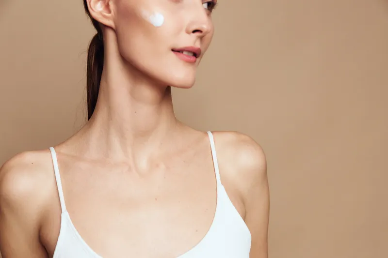 Young woman with clear skin wearing white top applies moisturizing cream on her cheek. Close-up on beige background. Concept of skincare, beauty, wellness, and self-care routine.