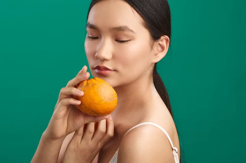 Asian woman holds an orange near her face in a clean studio setting, eyes softly closed and lips relaxed, radiating calm freshness and a subtle wellness vibe.