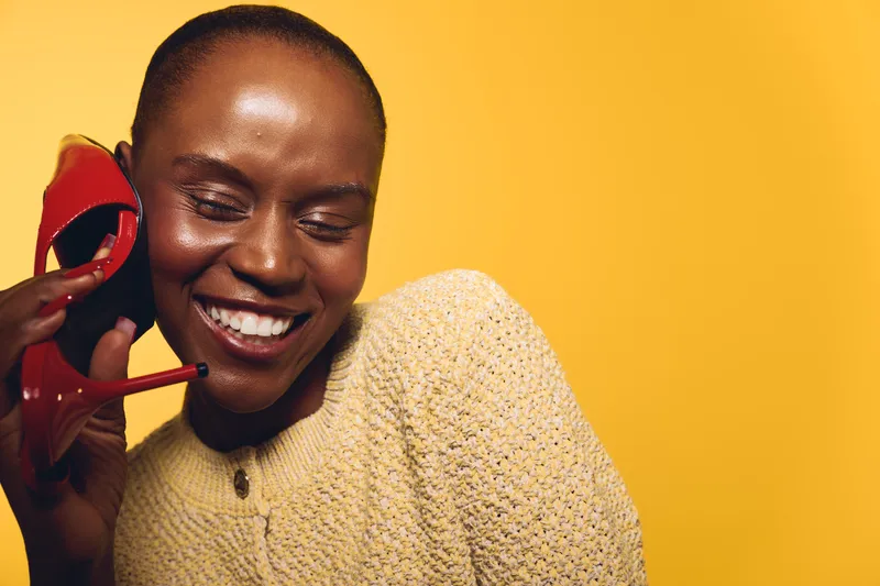 Smiling black woman with dark skin holds red high heel shoe near ear like phone, wearing beige knitted sweater. Bright yellow background creates cheerful mood and playful concept of fashion fun.