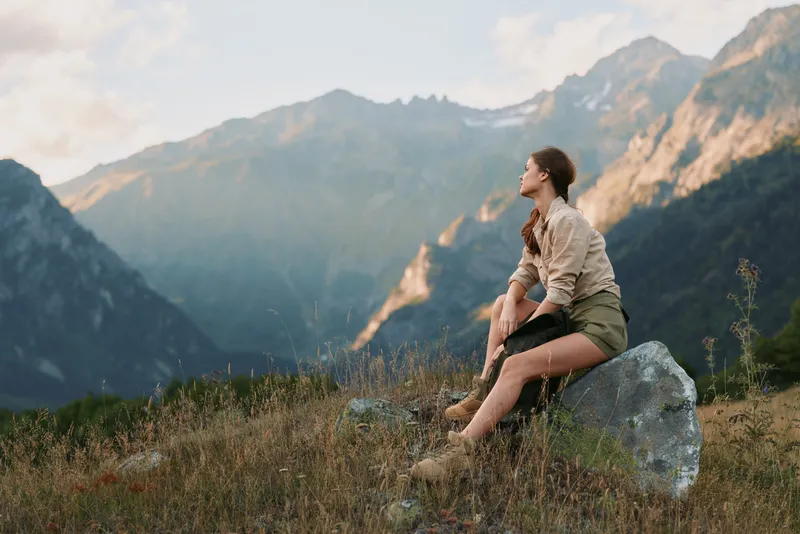 A young woman sits rock in a mountain landscape, gazing into the distance with a calm smile and relaxed posture amid a sunlit meadow.
