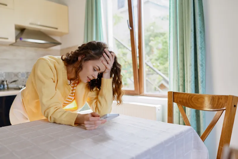 A distressed woman in a yellow cardigan sits at a dining table scrolling a tablet, head in hand, in a bright home kitchen, conveying burnout and loneliness.