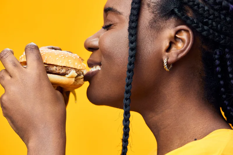 Smiling African woman enjoying a delicious hamburger, showcasing happiness against a vibrant yellow background, representing healthy eating and joyful moments