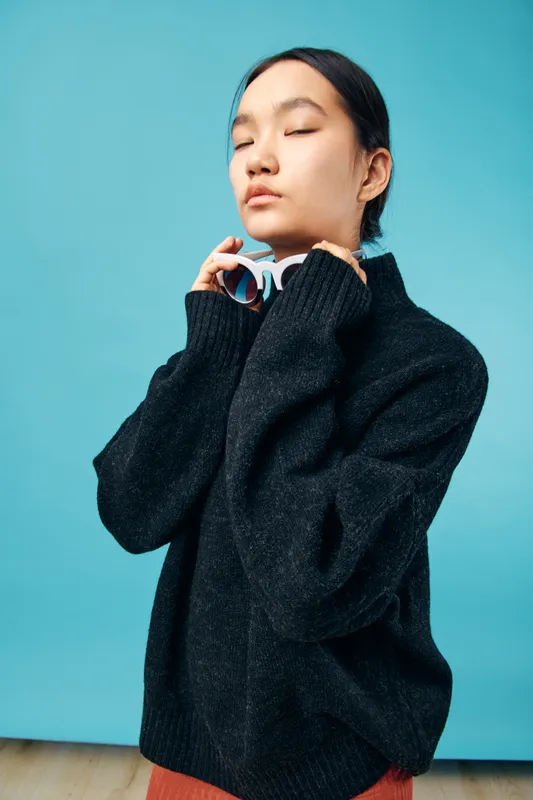 Young woman with closed eyes wearing oversized sweater holding glasses near face against blue background. Calm, thoughtful female with casual fashion and minimal studio setting portrait.