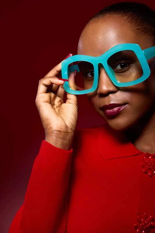 Close-up portrait of a confident African woman with dark skin wearing large blue glasses and a red sweater against a dark red background. Concept for fashion and style.