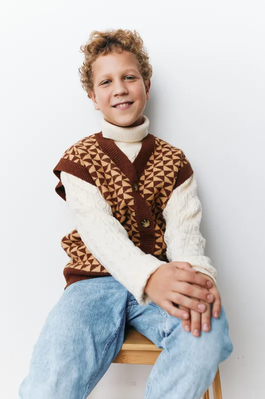 Authenticity and play theme blend as a cheerful child sits in a cozy knit vest, hinting at school routines, family moments, and everyday lifestyle in a warm, candid photograph.