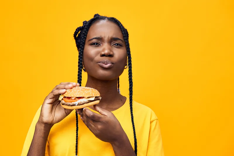 Young African woman holding a delicious burger with a playful expression against a vibrant yellow background, highlighting healthy eating and indulgence
