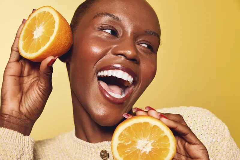 Happy smiling African woman with dark skin holding two orange halves near face on yellow background. Concept of fresh fruit, healthy lifestyle, vitamin, wellness, and natural beauty care.