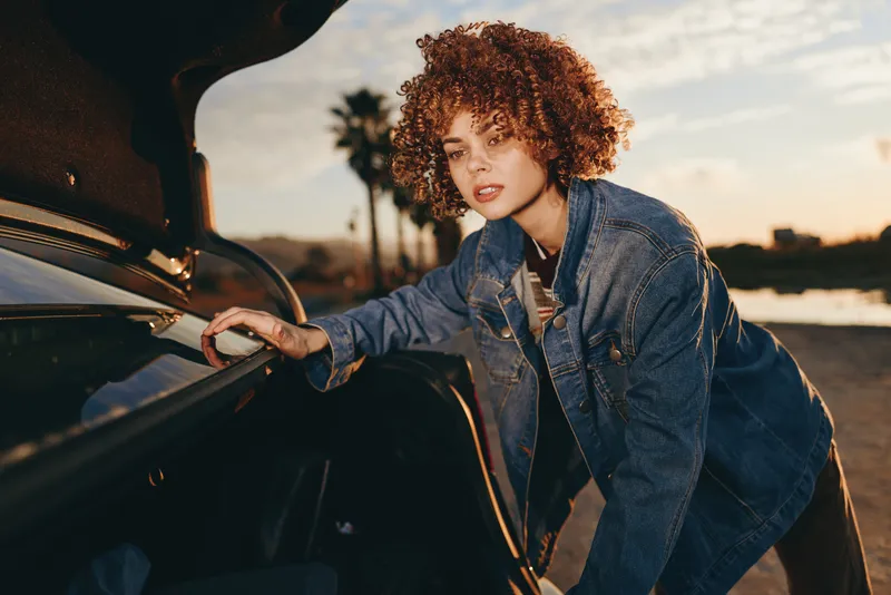 Woman lifestyle, curly hair, denim jacket, stands beside car door at sunset, one hand on vehicle, relaxed confident smile, outdoors road and street, urban golden light