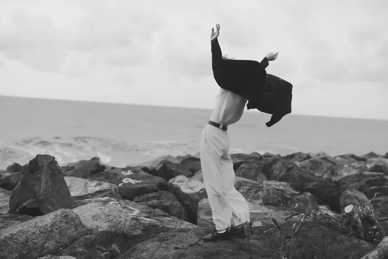 Businessman celebration outdoors by the coast with a jacket thrown into the air, rocks and ocean backdrop, conveying achievement, motion and freedom.
