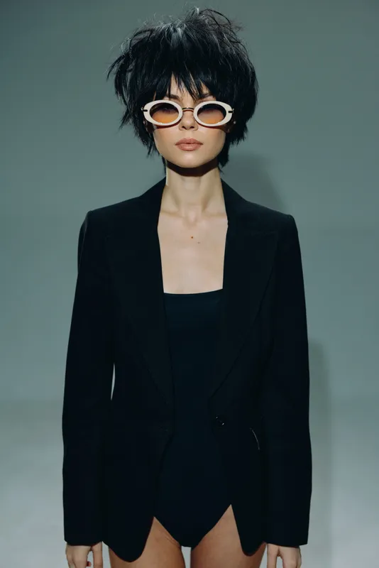 Stylish woman with short black hair wearing white sunglasses and black blazer against plain background