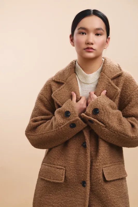 asian woman wearing a textured brown coat stands alone against a neutral backdrop, presenting a calm and thoughtful pose that conveys warmth, style, and confident poise.