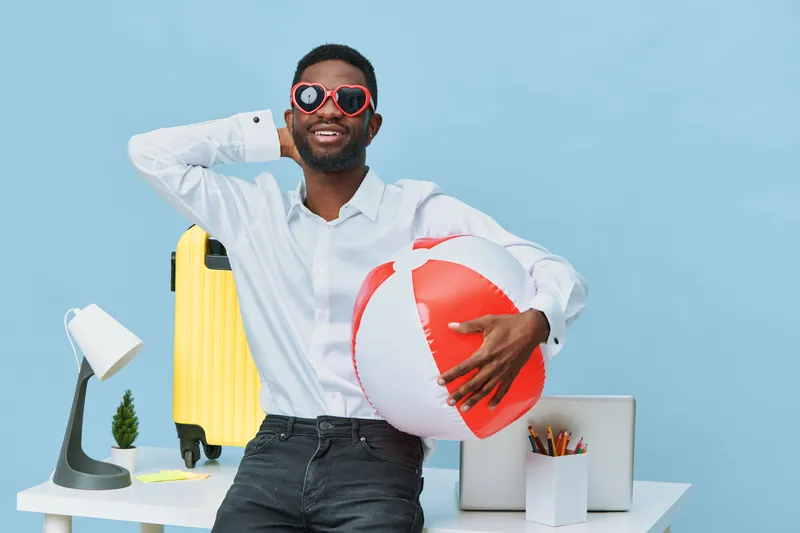 Happy cheerful man with sunglasses holding a red and white beach ball in bright summer indoor setting. Casual white shirt, black jeans, summer accessories, fun mood, modern work desk background with t