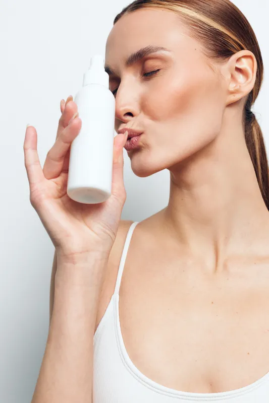 Young woman with clear skin holds white cosmetic spray bottle near face and kisses it with closed eyes isolated on white background in skincare concept.