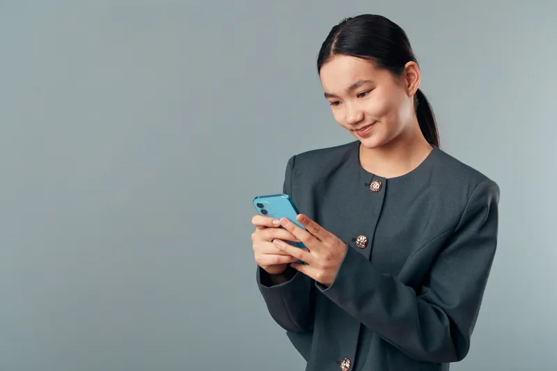 Young woman wearing dark blazer standing against grey background smiling and using blue smartphone for texting, social media or communication in casual modern style with neat hair.