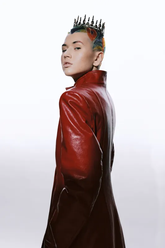 woman with short colorful hair wearing a red leather coat and black crown, posing confidently against a white background in studio lighting