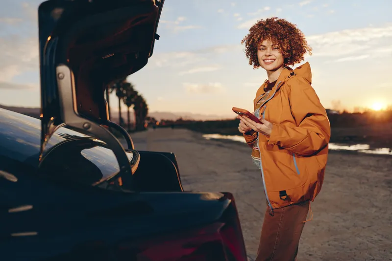 Woman lifestyle open car trunk at sunset, curly haired woman smiles and gestures while traveling on road outdoors, warm golden light, casual jacket, relaxed mood