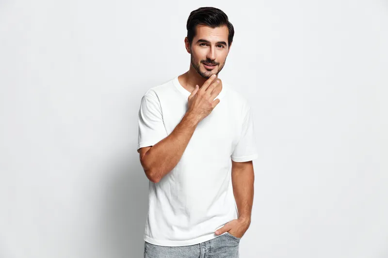 Handsome young man in a casual white t-shirt stands confidently with one hand on his chin, smiling at the camera against a plain solid background. People lifestyle concept.
