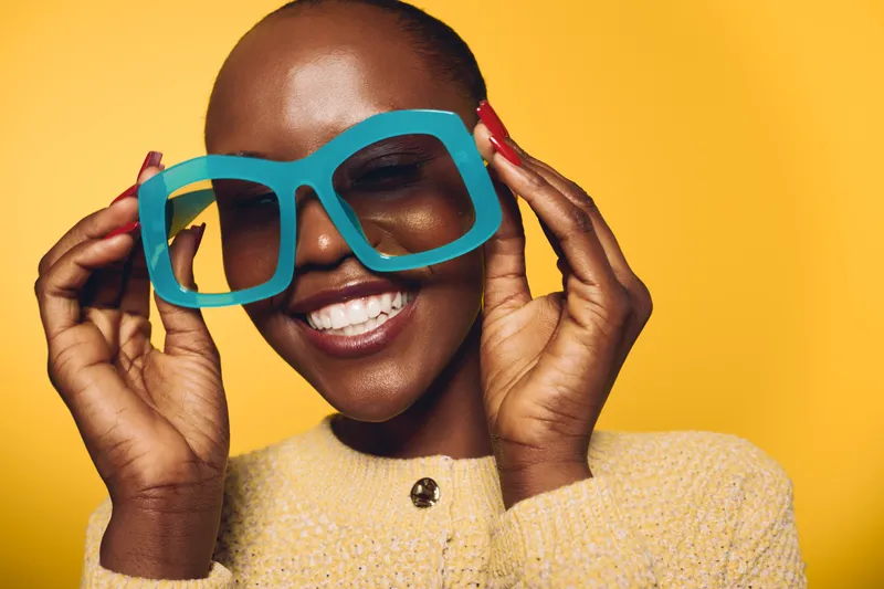 Smiling African woman with dark skin holds large blue glasses on yellow background. Happy female model in casual sweater expresses joy and confidence in bright studio lighting.