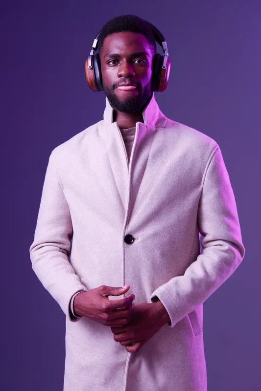Stylish young Black man wearing headphones and a beige coat, exuding confidence against a vibrant purple background, perfect for lifestyle and music themes