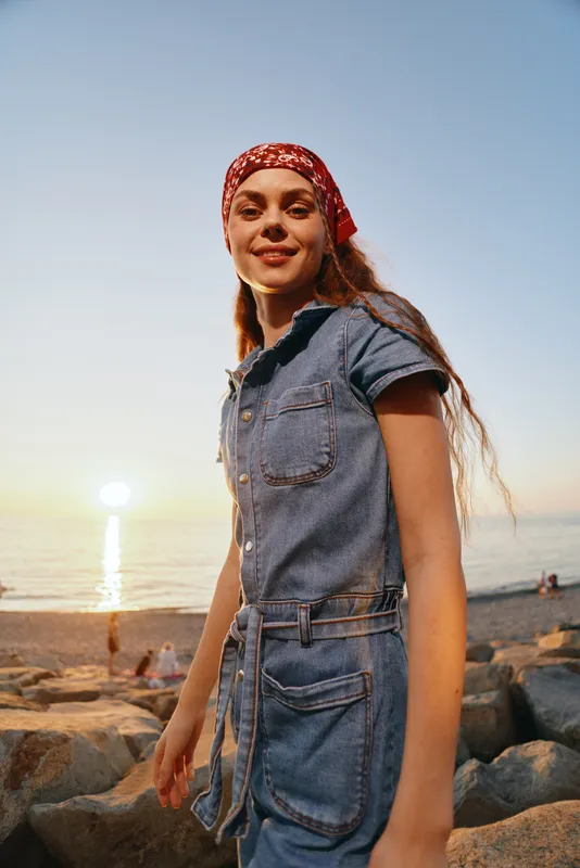 Lifestyle woman in modern boho-western denim street style smiling by the rocky beach at sunset with natural light and film color effects, casual and carefree mood.