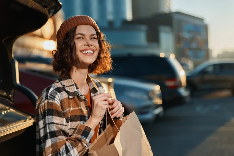 Woman smile shopping by car in parking lot during golden hour glow, candid lifestyle portrait with authenticity and mindful living, emotional storytelling of a cozy urban moment.