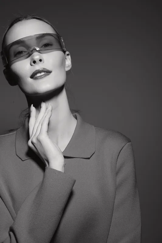 A young Caucasian woman with light skin wearing futuristic sunglasses and a textured jacket poses confidently in black and white. Concept of fashion and style.