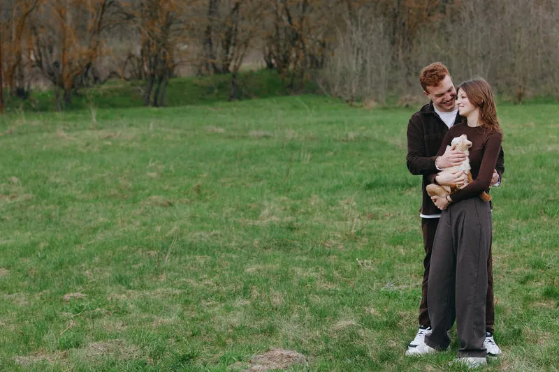 A couple stands in a green meadow, smiling as they cradle a young goat kid between them. Natural outdoor light highlights a warm, lifestyle moment of bonding.