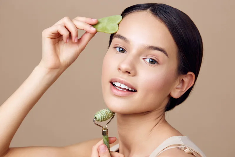 Woman using jade roller for facial massage, promoting skincare and natural beauty, showcasing a serene expression against a soft beige background