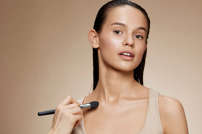 Young woman applying makeup with a brush on her shoulder, showcasing natural beauty and skincare concept with a smooth, warm background