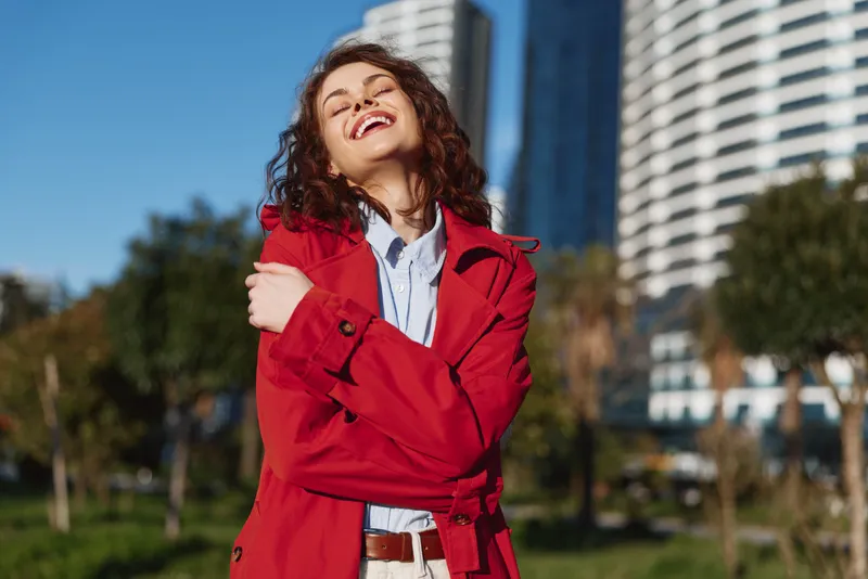 Smiling young woman with curly brunette hair wearing a red trench coat stands in a sunlit urban park, enjoying a carefree moment outdoors.