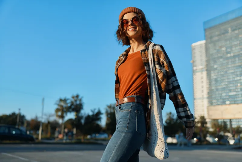 woman walking smile urban golden candid lifestyle authenticity: relaxed young woman strolling city plaza at golden hour glow, casual fashion in beanie and flannel, mindful living and emotional