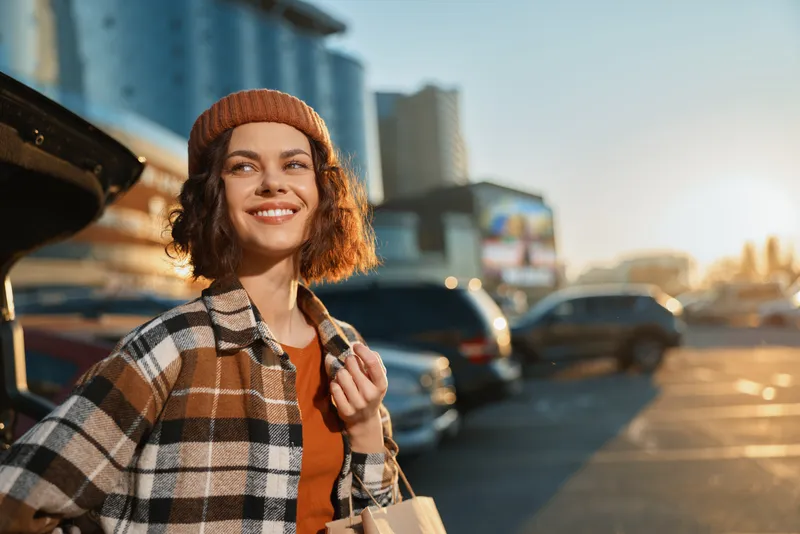 Woman smile urban parking candid lifestyle portrait at golden hour glow, beanie and autumn plaid coat, carrying shopping bag with authenticity, mindful living and emotional storytelling vibe.