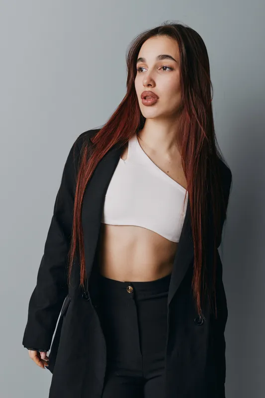 Young woman with long braided hair wearing black blazer and white crop top posing against gray background looking sideways with confident expression and stylish casual outfit.