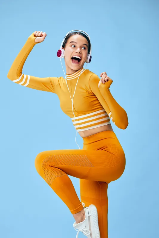 Fitness woman in bright orange athletic wear jumps with joy, dancing to music in a blue studio. Dynamic movement, energetic expression, headphones on, showcasing sport, fitness fashion and bold